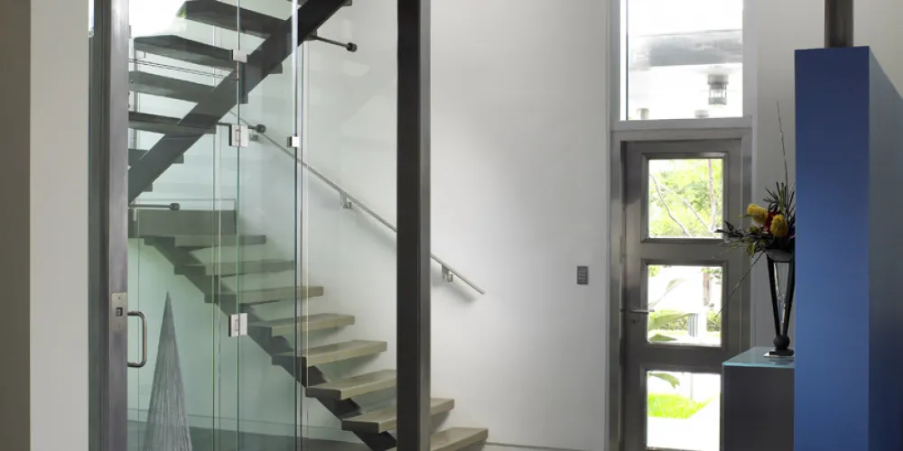This interior view showcases a modern staircase with an elegant glass elevator enclosure that adds depth and dimension to the space. The exposed steel beams highlight a blend of industrial design and modern minimalism. Natural light streams through a large, vertical window, emphasizing the concrete floor's clean lines and subtle textures.